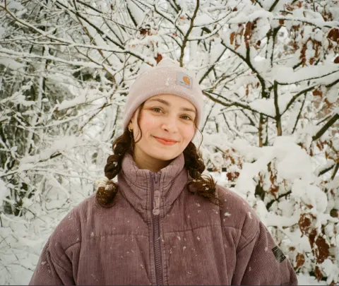 Person wearing a purple jacket and beige beanie, standing in a snowy forest with snow-covered branches in the background.