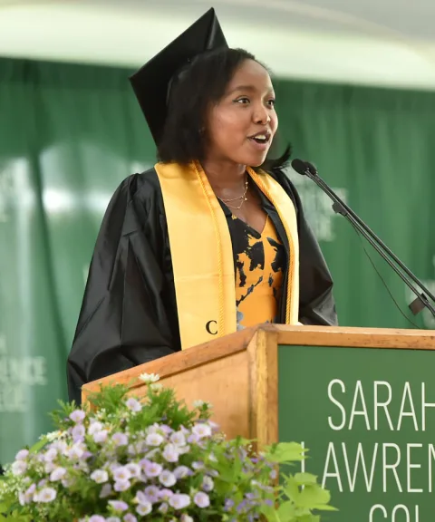 Person in graduation cap and gown speaks at a podium with "Sarah Lawrence College" sign, background features green curtain and flowers.