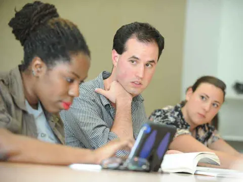 Three individuals are seated at a table, engaged in a discussion. One is typing on a laptop, with books in front of the others.