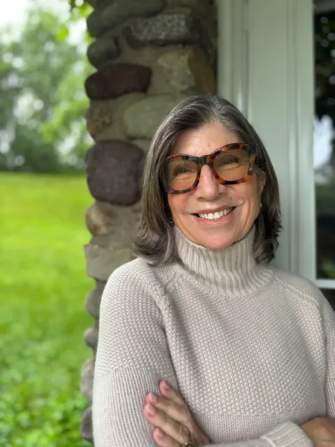 Person with glasses and gray hair smiling, wearing a light turtleneck sweater, standing in front of a stone wall and window. Greenery is visible outside.