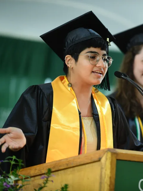 Graduate in cap and gown speaks at a podium, wearing glasses and a yellow stole, with a blurred background.
