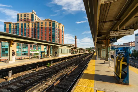 Train station platform with tracks leading toward a tall building and smokestack. Signs read "Yonkers" and direct to Croton-Harmon and Poughkeepsie.