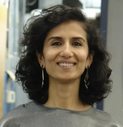 Person with curly hair, smiling, wearing a gray top and dangling earrings, standing indoors.