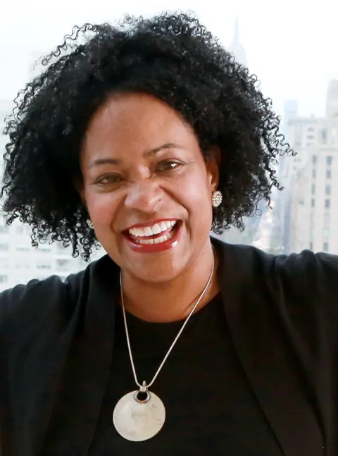 Smiling individual with curly hair, wearing a dark outfit and a round pendant necklace, standing in front of a city skyline.