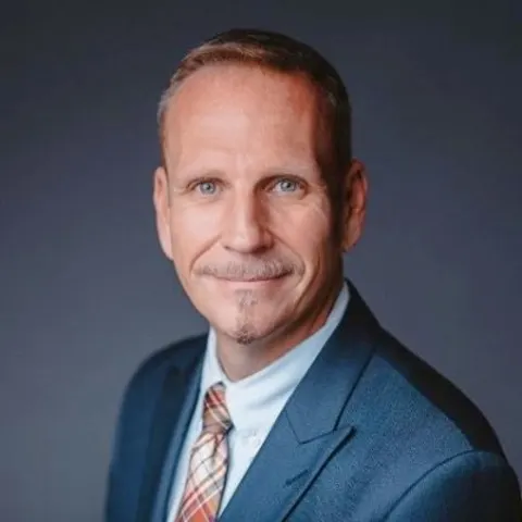 Person in a suit with a plaid tie, smiling against a dark background.