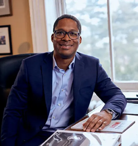 Person wearing glasses and a blue suit is sitting at a desk in an office with framed pictures and a window in the background.