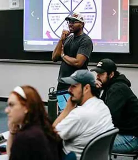 Classroom setting with an individual standing in front of a presentation screen, while others sit at tables, focusing on the presentation.