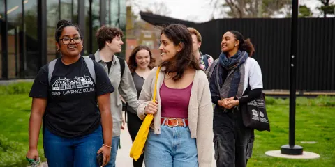 A group of people walking and chatting on a campus path, with one wearing a "Sarah Lawrence College" shirt, near a modern building.