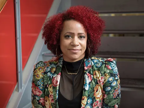 Person with curly red hair and floral jacket sits on stairs, smiling slightly, against a red and gray background.