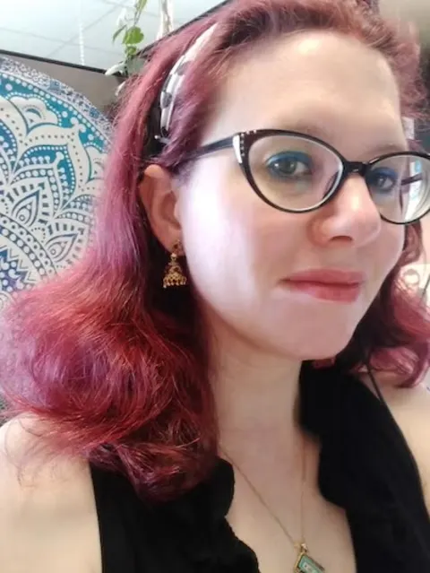 Person with red hair, glasses, a headband, and earrings, wearing a black top. A patterned tapestry is in the background.