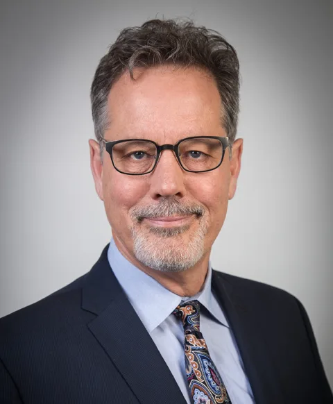 Person with glasses and facial hair, wearing a suit with a paisley tie, against a plain background.