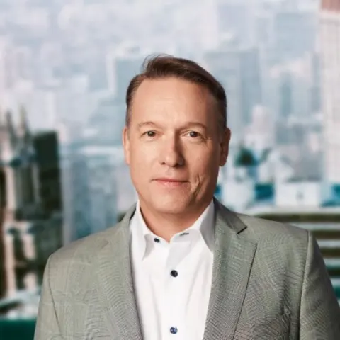 Person wearing a gray suit and white shirt, standing in front of a blurred cityscape background.
