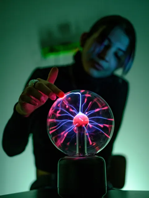 Person touching a plasma globe with glowing pink and blue electric streams inside a dimly lit room.