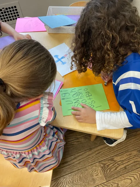 Two kids sit at a table, drawing on colorful paper. A basket with more paper is in the background.