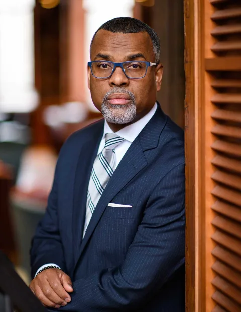 Person in a suit with striped tie, wearing glasses, leans against a wooden structure in an indoor setting.