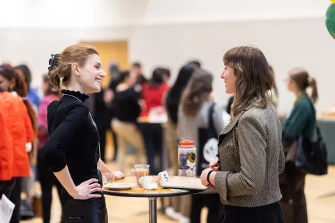 Two people engaged in conversation at a networking event, standing by a table with drinks and snacks. Groups are mingling in the background.