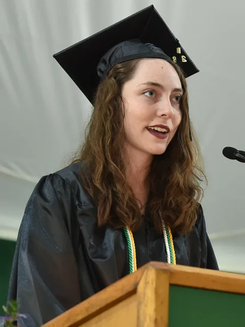 Person in graduation cap and gown speaking at a podium during a ceremony.