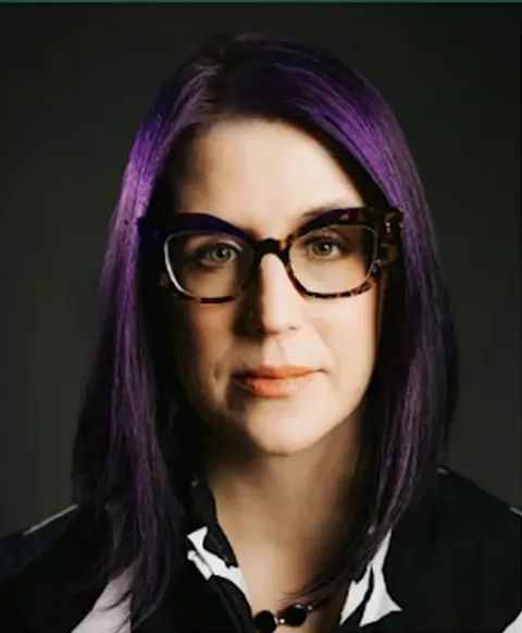 Person with long purple hair, wearing glasses and a black and white shirt, looking at the camera against a dark background.