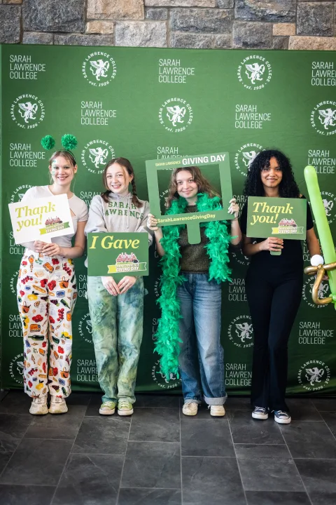 Four people holding signs for Sarah Lawrence College Giving Day pose in front of a college-branded backdrop.