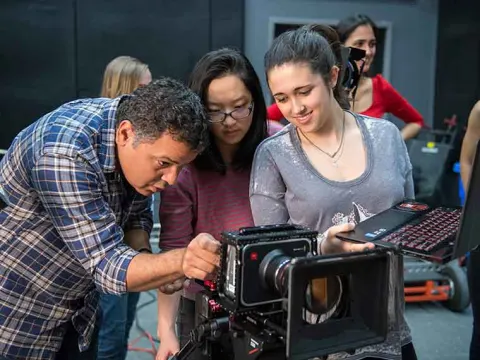 People collaborate around a camera on set, one holds a laptop while another adjusts settings.