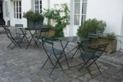Outdoor patio with metal tables and chairs on a cobblestone surface, surrounded by potted plants and white building walls.