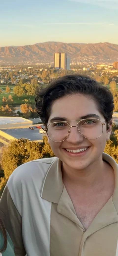 Person smiling with glasses, short dark hair, and a collared shirt. Background shows trees and a distant mountain range.