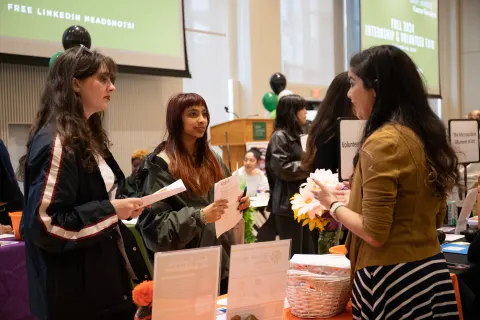 three people conversing at a conference booth; others in the background