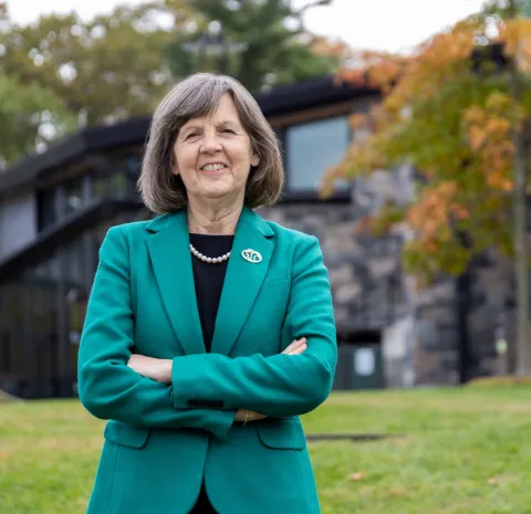 Person in a green blazer with arms crossed, standing in front of a modern building with autumn trees in the background.