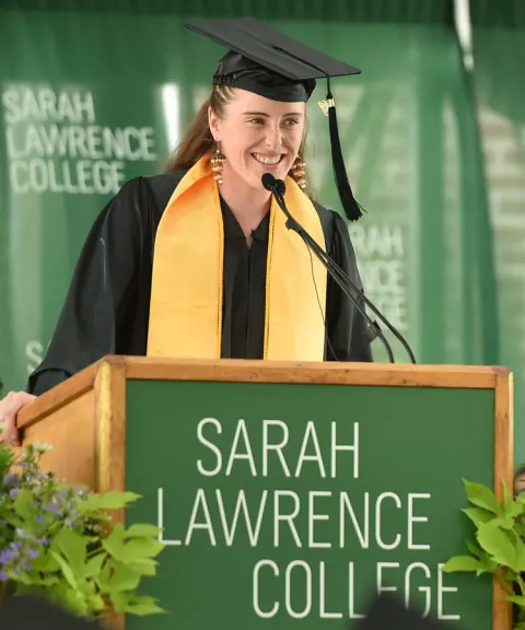 Person in graduation attire speaks at podium with "Sarah Lawrence College" sign.