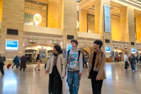 Three people walk through a large, brightly lit train station with a high ceiling and ornate columns, with other people in the background.
