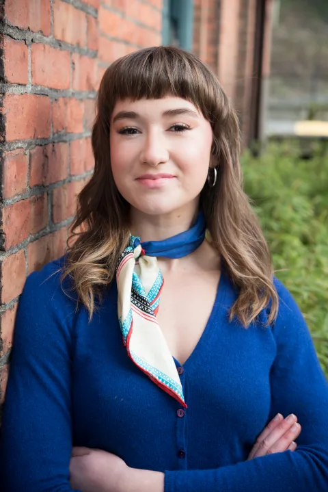 Person with long hair and a fringe, wearing a blue top and patterned scarf, stands against a brick wall with folded arms.