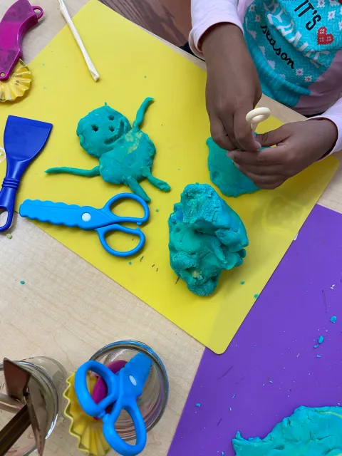Child shaping blue play dough on a yellow mat with blue scissors, a clay figure, and tools nearby.