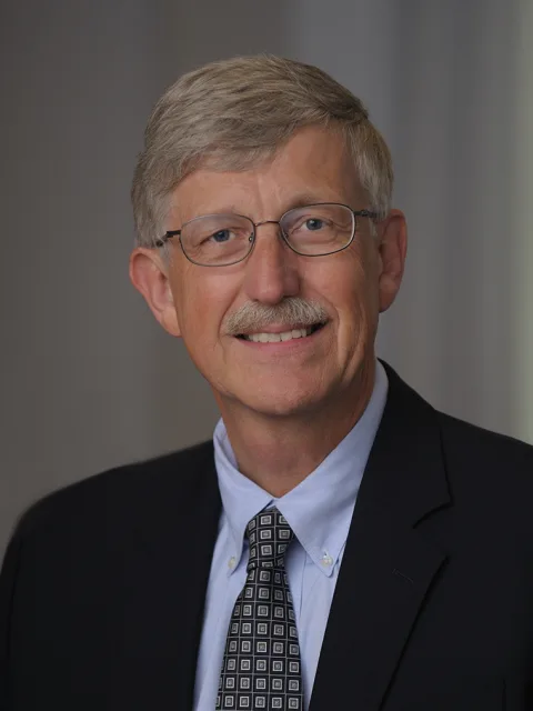 Person with glasses, light hair, and mustache, wearing a suit, light blue shirt, and patterned tie, smiling against a neutral background.