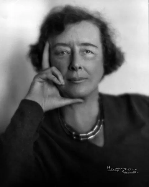 Person in a dark top, resting chin on hand. Necklaces visible, with a thoughtful expression against a neutral background.