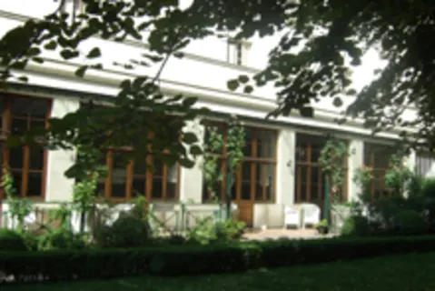 A building with large wooden-framed windows is surrounded by lush greenery and a manicured garden, partially shaded by tree branches.