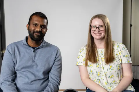 Two people sitting and smiling at the camera, one wearing a blue sweater and the other wearing a floral shirt, against a plain backdrop.