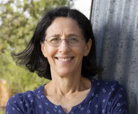 Person with glasses smiling outdoors, wearing a blue patterned shirt, standing near a corrugated metal surface with trees in the background.