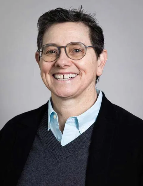 Smiling person with short hair, wearing glasses, a light blue shirt, grey sweater, and black jacket against a neutral background.