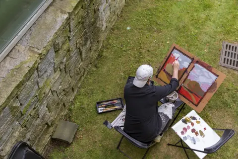 Person painting outdoors, seated on a lawn near a stone wall. Art supplies are scattered around, with a landscape painting taking shape.