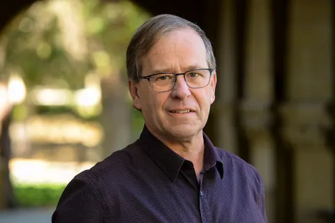 Person with glasses and short hair wearing a dark shirt, standing in an outdoor setting with blurred trees and arches in the background.