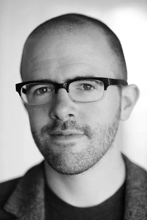 Person with glasses and a beard, looking at the camera. Black and white photo with a neutral expression.