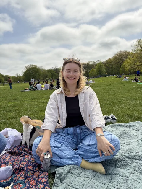 Person sitting on a blanket in a park, smiling, holding a drink can, with a bag nearby and others relaxing in the background.