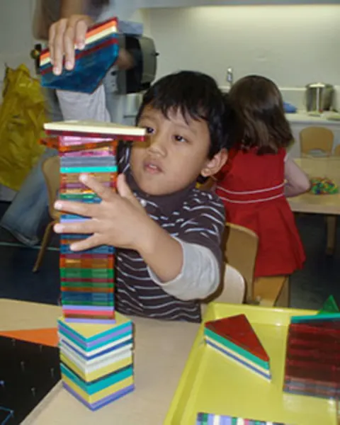 Child stacking colorful geometric tiles at a table, focused on balancing them. Another child is in the background.