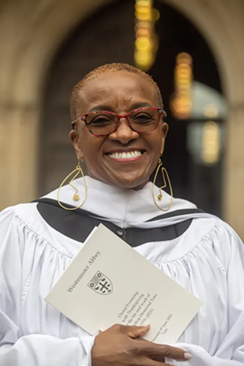 Smiling individual in ceremonial robe holding a Westminster Abbey program, standing in front of an arched entrance with blurred lights behind.