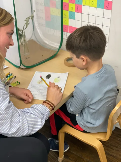 Person showing a drawing to a child at a desk with crayons, paper, and a chart in the background.