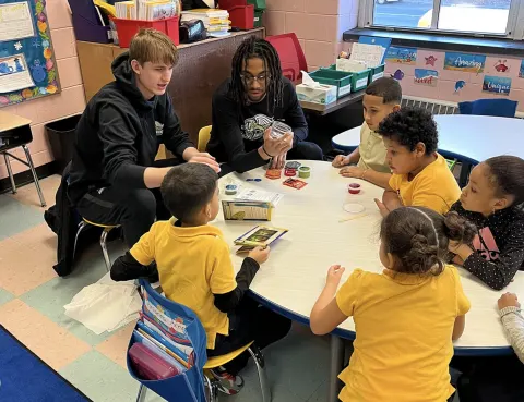 Two individuals engage with children around a table filled with craft materials in a classroom setting.