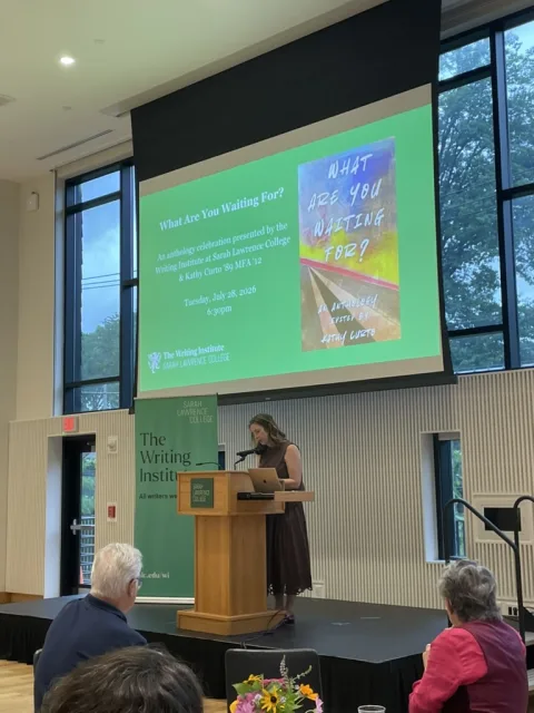 Person standing at a podium speaking at an event with a screen displaying book details. Audience members are seated in front.