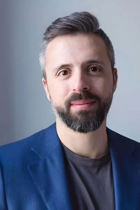 Person with short gray hair and beard, wearing a blue blazer and gray shirt, smiling slightly against a neutral background.