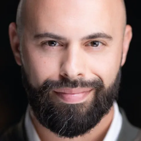 Person with a bald head and a thick, dark beard smiling softly, shown in close-up with a blurred dark background.
