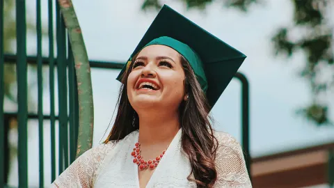 Person in green graduation cap smiling, wearing a lace top and red necklace, with a blurred outdoor background.